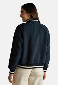 Tumesinise fliistekk, millel on soonik kaelus ja mansetid, valgete triipudega. Kasti kujuline disain pehme tekstuuriga, nähtavad õmbluse detailid.