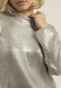 Pull en argent, à côtes, avec un col haut et des manches longues. Le tissu a une texture brillante et réfléchissante.