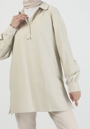Vrouw die beige tuniek met lange mouwen, knoopmanchetten en gedeeltelijke knoopsluiting draagt over een wit hemd en lichte broek, staand.