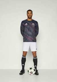 Equipación de fútbol que presenta una camiseta de manga larga con un patrón oscuro y pantalones cortos blancos, calcetas negras con tres franjas y botas de fútbol negras.