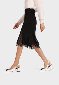 Gonna nera di satin con orlo in pizzo e spacco laterale; abbinata a scarpe con tacco a blocco bianche con dettagli neri e punta aperta.