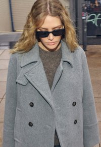 Gray double-breasted coat made of wool blend, featuring large buttons and notched lapels, worn over a dark sweater, paired with sunglasses.