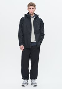 Jachet�ă impermeabilă neagră cu glugă, pulover gri și pantaloni de trening negri cu dungi albe pe laterale. Purtați cu pantofi sport gri.