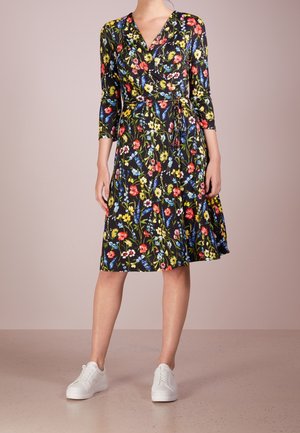 Femme portant une robe portefeuille noire longueur genou avec un motif floral coloré et des baskets blanches, debout devant un fond clair uni.