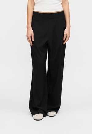 Fekete, bő szárú nadrág sima textúrával és elasztikus derékpánttal, világosszürke bebújós cipővel párosítva. Egyéb jelentős részlet nincs.