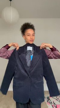 Osoba o kręconych włosach trzyma duży czarny blezer z niebieską metką w minimalistycznym pokoju z papierową latarnią i wzorzystym dywanem.