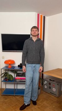 Junger Mann in grauem Kapuzenpullover und hellen Jeans steht im Wohnzimmer mit gestreifter Wand, blauem Regal, orangefarbener Lampe und Koffer mit Aufklebern.