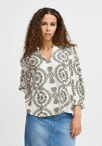 Hvid mønstret bluse med sorte blomsterdesigns, med en V-udskæring, lange puffærmer og en afslappet pasform. Sammen med en denimnederdel.