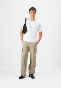 T-shirt en coton blanc avec un logo noir, pantalon cargo beige avec plusieurs poches, et baskets noires avec des accents blancs.
