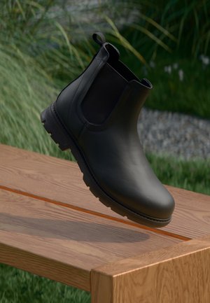 Black leather Chelsea boot with elastic sides, rounded toe, and textured sole, hovering above a wooden surface with greenery in the background.