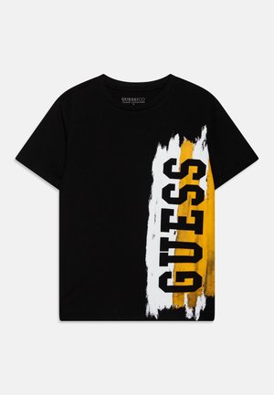Czarny t-shirt z krótkim rękawem z pionowym wzorem białych i żółtych pociągnięć pędzla oraz wyraźnym czarnym napisem "GUESS" po prawej stronie.