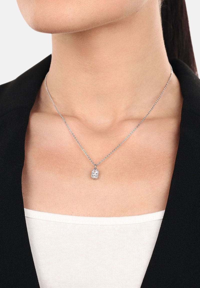 Collana in argento con pendente ovale e pietra trasparente, indossata sopra un top neutro e una giacca nera.