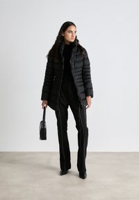 Zwarte pufferjas met een hoge kraag en knoopsluiting, gedragen met zwarte pantalons met zijnaad. Model houdt een kleine zwarte handtas vast.