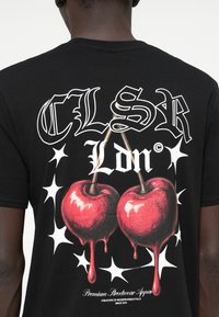 Μαύρη βαμβακερή μπλούζα τύπου t-shirt με γραφικό τύπωμα από κόκκινες κερασιές με λεπτομέρειες που στάζουν, περιτριγυρισμένες από λευκά αστέρια και έντονο κείμενο, στο πίσω μέρος.