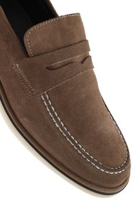 Mocassin en daim marron avec un bout mocassin cousu, présentant une bande texturée sur le dessus et un accent de semelle en caoutchouc blanc.
