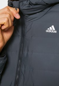 Giubbotto imbottito grigio scuro con colletto alto, chiusura con zip verticale e logo Adidas ricamato in bianco sul lato sinistro del petto.