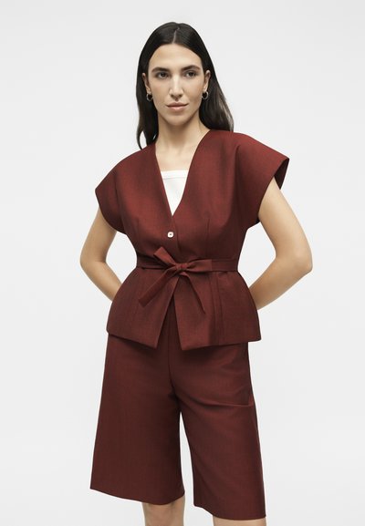 Γυναίκα που φοράει ένα σκουριάς κοντομάνικο γιλέκο με ζώνη και κουμπί, ταιριαστά σορτς μέχρι το γόνατο και λευκό τοπ από μέσα.