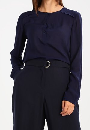 Vrouw draagt een marineblauwe blouse met lange mouwen en knopen, en een marineblauwe broek met hoge taille en een zwarte riem met een zilveren gesp.