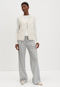 Strukturierte cremeweiße Weste mit beigem Besatz, getragen über einem weißen Oberteil und kombiniert mit grauen, weit geschnittenen Hosen. Auffällig ist der Reißverschluss der Weste.