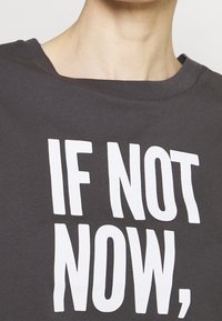 Camiseta de algodón gris carbón con un escote amplio y mangas cortas, que presenta un gran texto blanco que dice: "SI NO AHORA,".