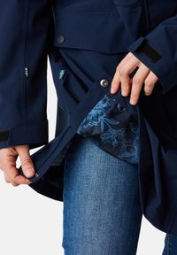 Giacca impermeabile navy con interno foderato a fiori, chiusura a scatti, polsini regolabili e tag di branding discreto visibile sulla manica.