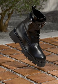 Czarne skórzane botki w kostkę z wytrzymałą podeszwą, sznurowanym przodem i regulowanym paskiem z klamrą. Gładka tekstura z błyszczącym wykończeniem.