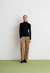 Kvinde står op ad en hvid væg iført en sort turtleneck, beige corduroybukser og sorte sko på et grønt gulv.