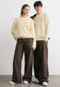 Deux mannequins portant des pulls crème assortis avec des logos verts, des pantalons larges marron et des baskets blanches, debout devant un fond blanc uni.
