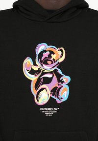 Felpa nera con cappuccio caratterizzata da una grafica colorata di un orso con occhi a forma di X. Il testo sottostante recita "CLOSURE LDN" e l'anno di fondazione. Tessuto morbido.