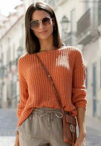Orange gestrickter Pullover mit lockerer Passform, langen Ärmeln und einem strukturierten Muster. Kombiniert mit hellgrauen Hosen und einer kleinen braunen Umhängetasche.