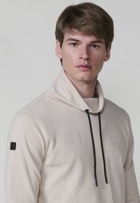 Giovane uomo con capelli castano chiaro che indossa un top beige a maniche lunghe con un collo alto e lacci neri su uno sfondo neutro.