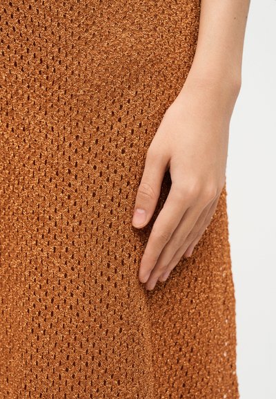 A jobb kéz finoman pihen egy texturált, rozsdaszínű, apró lyukakkal tűzdelt kötött anyagon.