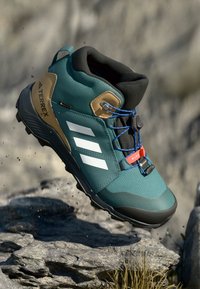 Zeleni vodootporni planinarski čizme s crnim i smeškastim akcentima, koja ima teksturiranu gornju površinu, tri bele pruge i plave vezice.