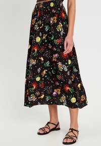 Jupe midi noire à motifs floraux avec une taille smockée, présentant des motifs colorés de soleils et d'animaux. Portée avec des sandales noires à brides.