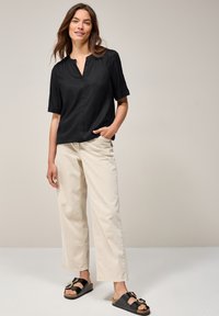 Blouse noire à manches courtes avec un col mandarin, associée à un pantalon large beige et des sandales noires à glisser avec des accents dorés.