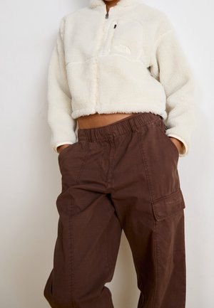 Osoba oblečená v krémově zbarvené fleecové bundě se zipem a tmavě hnědých pracovních kalhotách s elastickým páskem, stojící s rukama v kapsách.