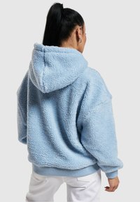 Lyseblå, fluffy fleece hettegenser med stor hette, ribbestrikkede mansjetter og kant. Stoffet har en teksturert, myk utseende.