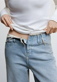 Jasnoniebieskie dżinsy z odsłoniętymi szwami i postrzępionym pasem. Dopasowana, biała, długorękawowa bluzka częściowo zasłania pas.