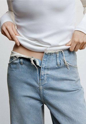 Světlé modré džíny z denimu s odhaleným švem a roztrhaným pasem. Přiléhavý bílý top s dlouhým rukávem částečně zakrývá pas.