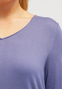 Sweter z dekoltem w serek w kolorze lawendowym, wykonany z gładkiego dzianinowego materiału o delikatnej fakturze i krótkich rękawach. Znajduje się w nim wyraźna prostota.
