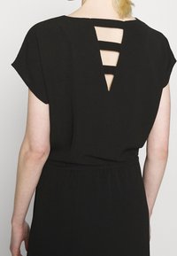 Vista trasera de una persona con un vestido negro de mangas cortas y un corte trapezoidal en la parte superior de la espalda, con tres tiras horizontales.
