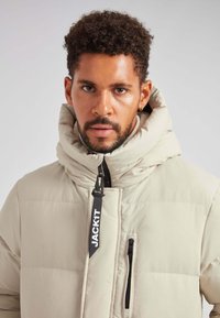 Giacca puffer beige chiaro con colletto alto e cappuccio oversize. Presenta una chiusura con zip e una tasca frontale sul petto con hardware nero.
