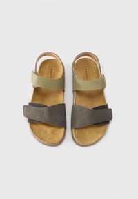 Δύο επίπεδες σαγιονάρες με λουριά σε ελιά πράσινο και σκούρο γκρι. Οι εσωτερικές σόλες είναι καφέ με απαλή,  υφασμάτινη υφή.