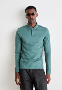 Cămașă polo cu mânecă lungă de culoare teal, cu un guler butonat, având un logo subtil brodat, combinată cu pantaloni din denim închis.