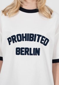 T-shirt blanc avec bordure noire sur le col et les manches, arborant de grandes lettres noires texturées formant le texte "PROHIBITED BERLIN".