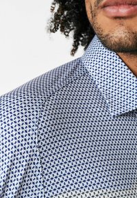 Hemd mit Knopfleiste in einem blau-weißen geometrischen Muster, mit klassischem Kragen und strukturiertem Stoff. Nahaufnahme des Ausschnitts und der Schulter.