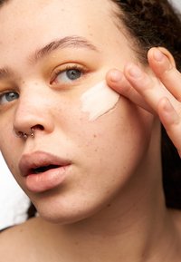 Personne jeune avec un piercing au septum appliquant de la crème sur la joue avec les doigts, gros plan montrant la texture de la peau et de légères taches de rousseur.