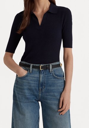 Kvinde iført en sort kortærmet ribbet poloshirt, der er stoppet ned i højttaljede blå jeans med et sort bælte.
