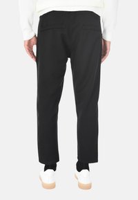 Vista posteriore di una persona che indossa pantaloni neri a gamba dritta, sneakers bianche e una maglietta bianca a maniche lunghe, in piedi su uno sfondo neutro.