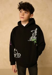 Garçon aux cheveux bouclés portant un sweat à capuche noir avec des squelettes blancs à vélo et du texte vert, se tenant devant un fond beige.